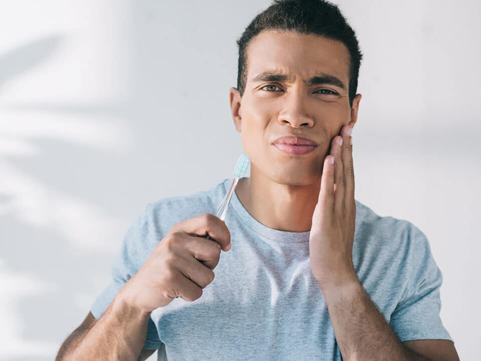 young man holds a toothbrush and his cheek as he's experiencing one of the 9 common causes of tooth pain