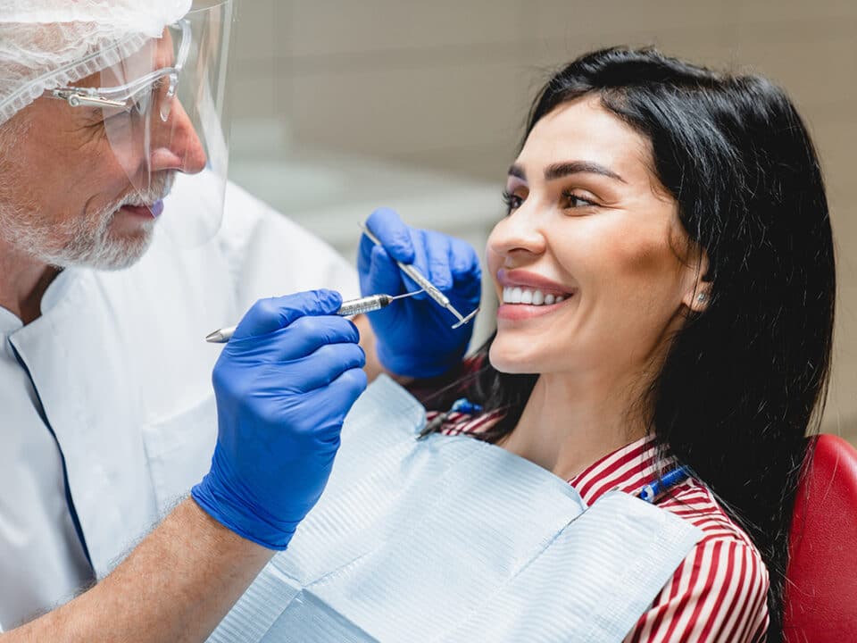dentist with a mirror and pick tool in hand inspects a woman's veneers after she asked can dental veneers be reshaped?