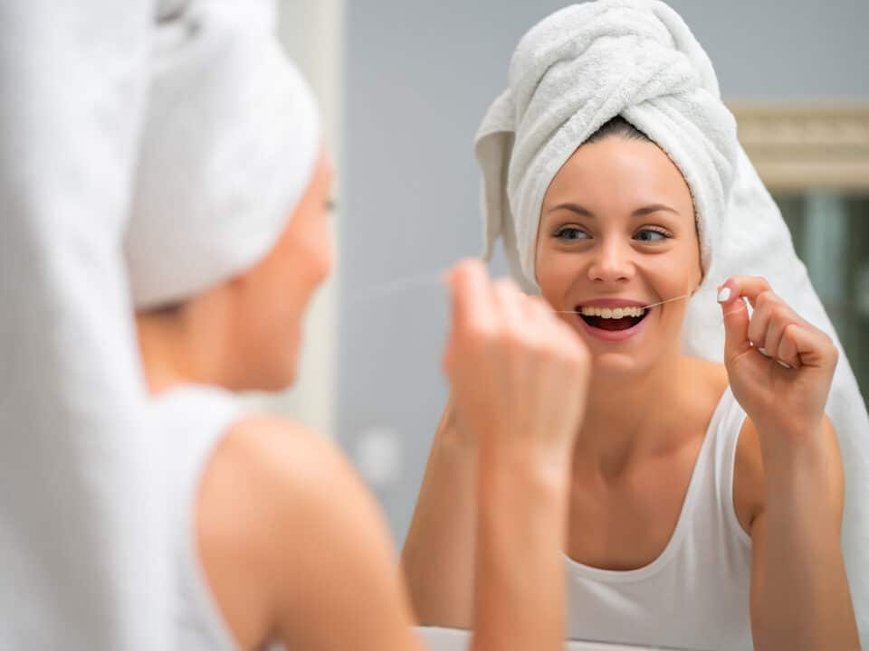 woman with her hair wrapped in a towel flosses her teeth in a mirror