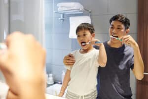 father and son brush their teeth together in the mirror