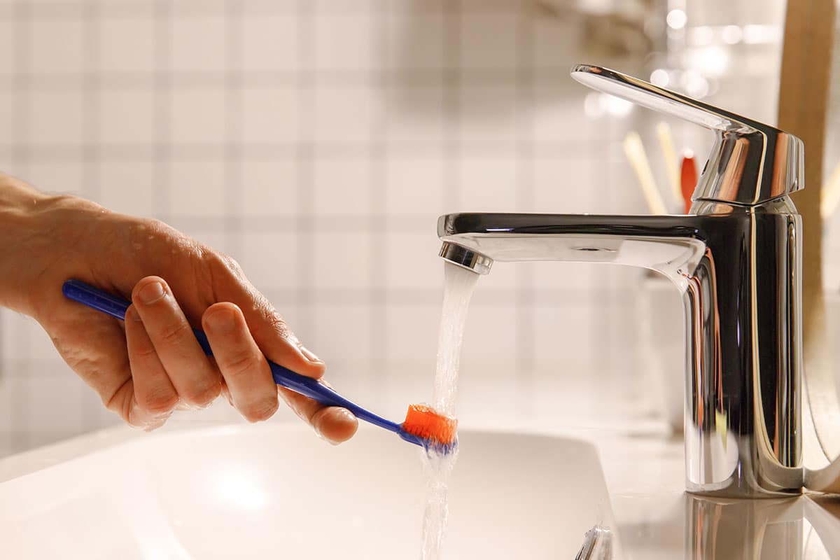 man runs his toothbrush under hot water as part of his how to brush your teeth properly routine