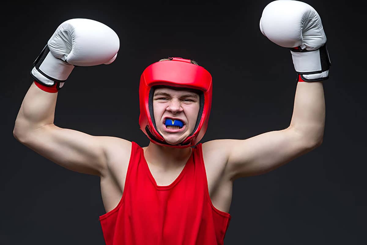 a male boxer in red shirt and helmet puts his fist in the air as he shows off a sports mouth guard that is protecting his teeth