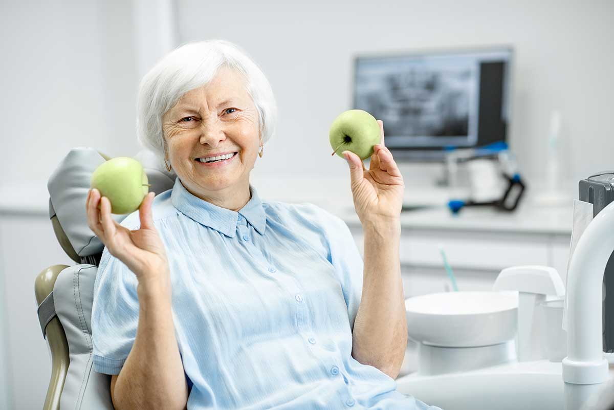 elderly woman holds up two apples which she can now eat due to one of the many advantages of dental implants