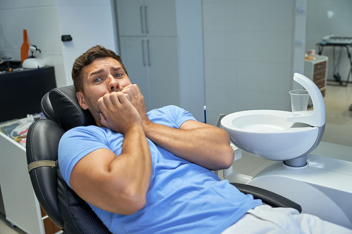 man sitting hte dentist chair puts his hands up to his face as he suffers from dental anxiety
