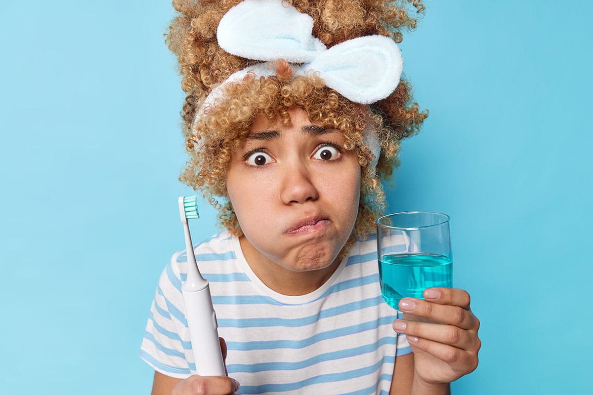 photo of a woman holding a toothbrush and some mouthwash as she looks confused about the benefits of using mouthwash