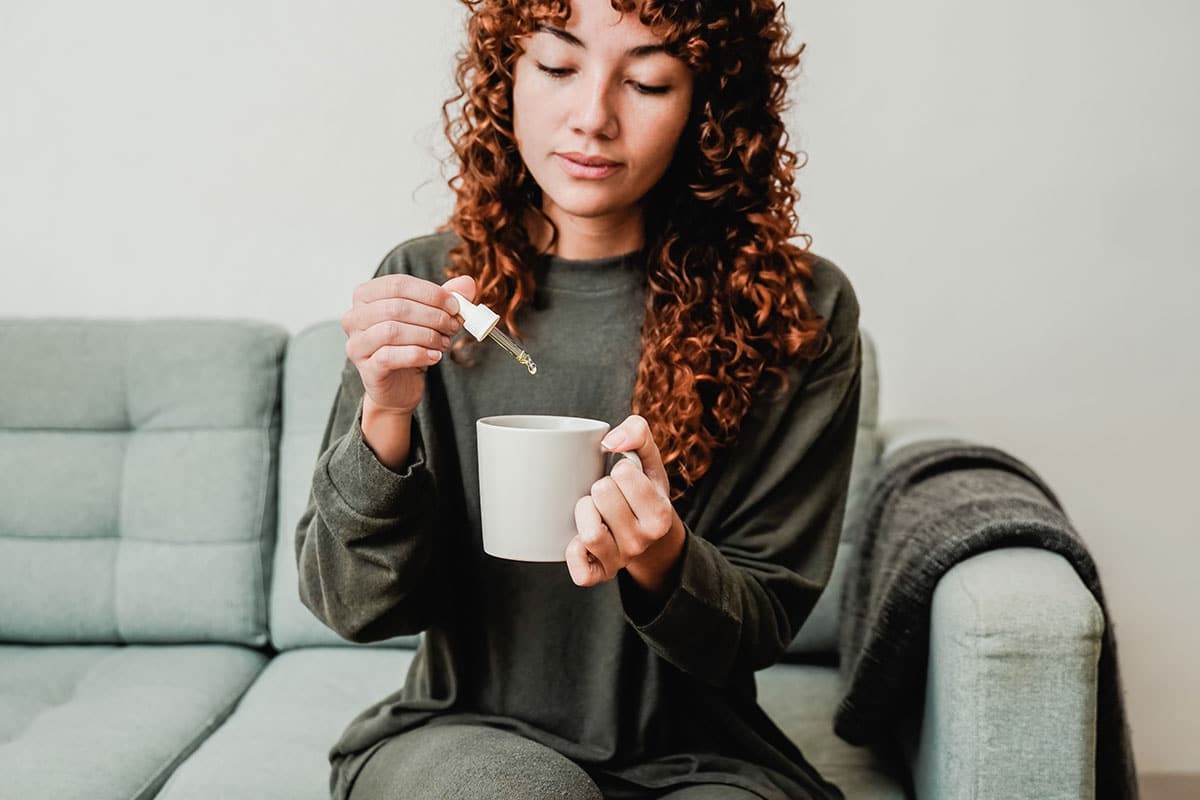 red haired woman in a green sweater uses an eye dropper to add some cbd oil drops to her tea