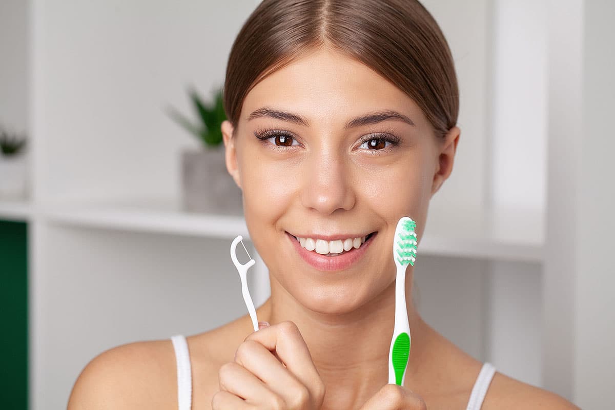 young woman holds up a floss pick and a toothbrush wondering is flossing is better than brushing