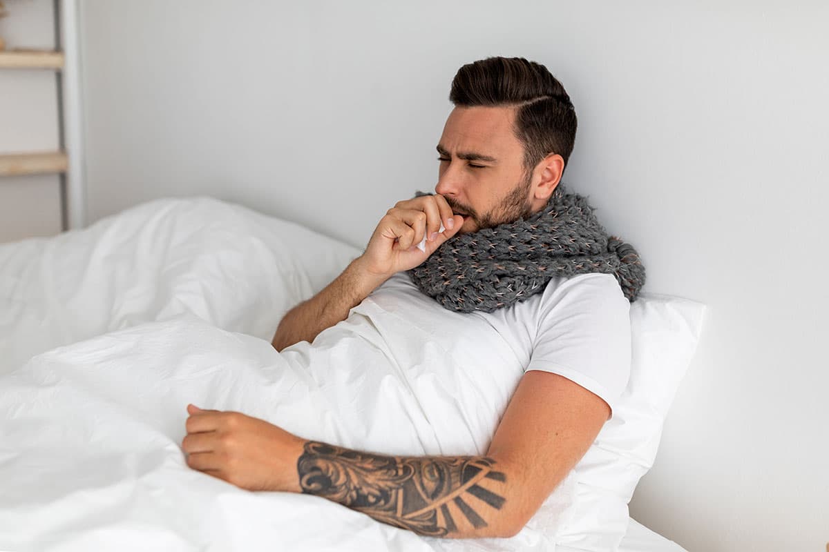 man sits in bed coughing with a shall wrapped around his neck