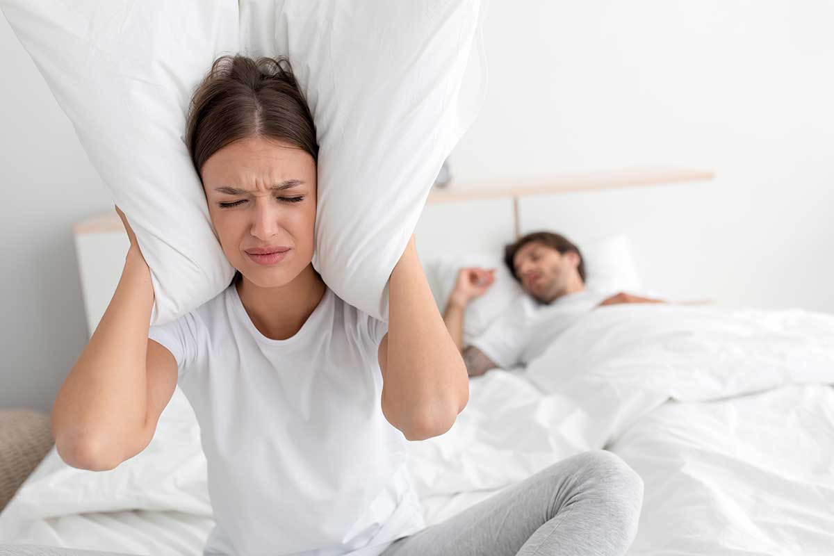 woman blocking her ears with a pillow because her partner is exhibiting the snoring symptom of sleep apnea