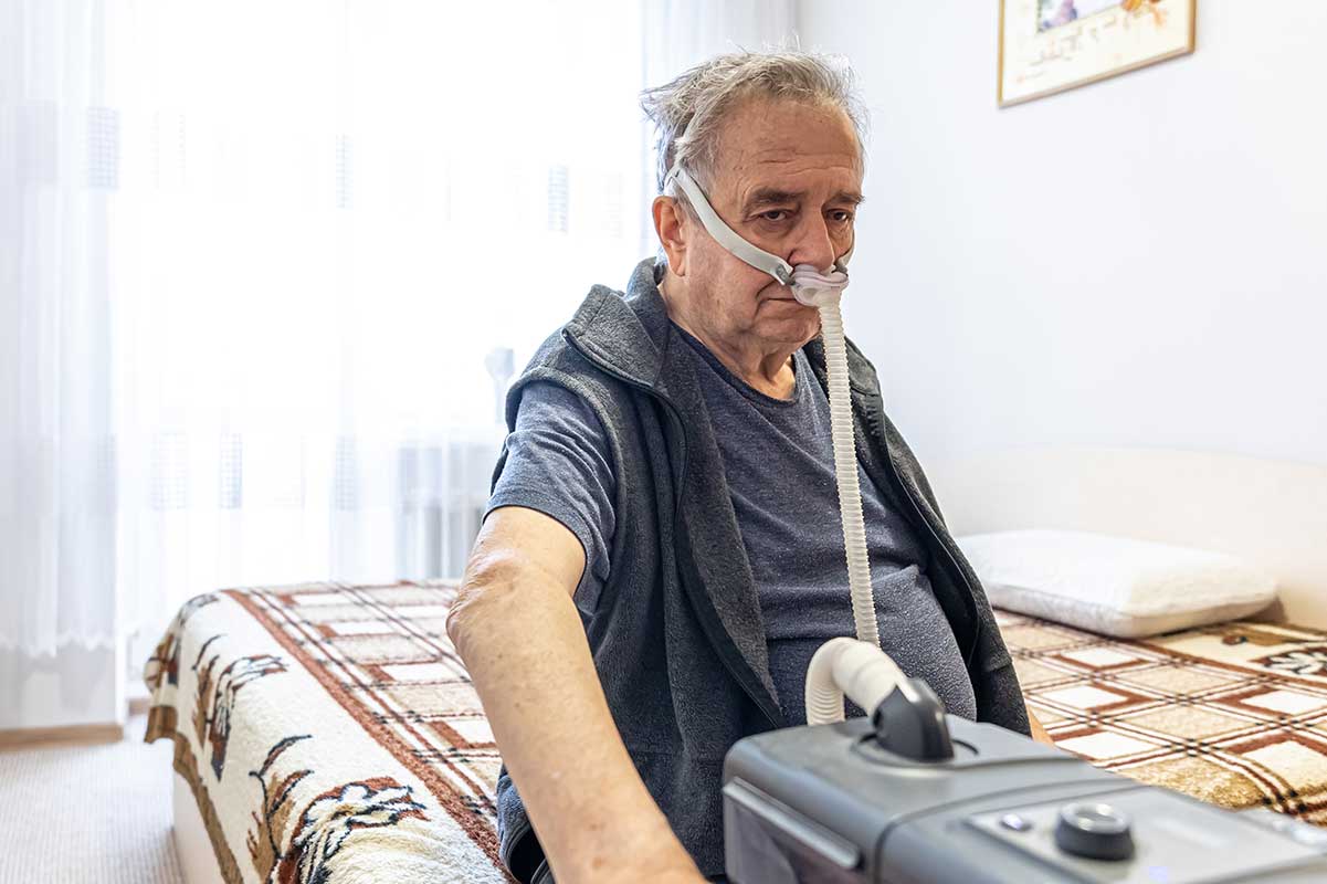 senior man sits on his bed looking sad because he's chained to his cpap machine from untreated sleep apnea