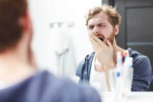 man yawning in the mirror about to brush his teeth after reading our article about tooth care for lazy people