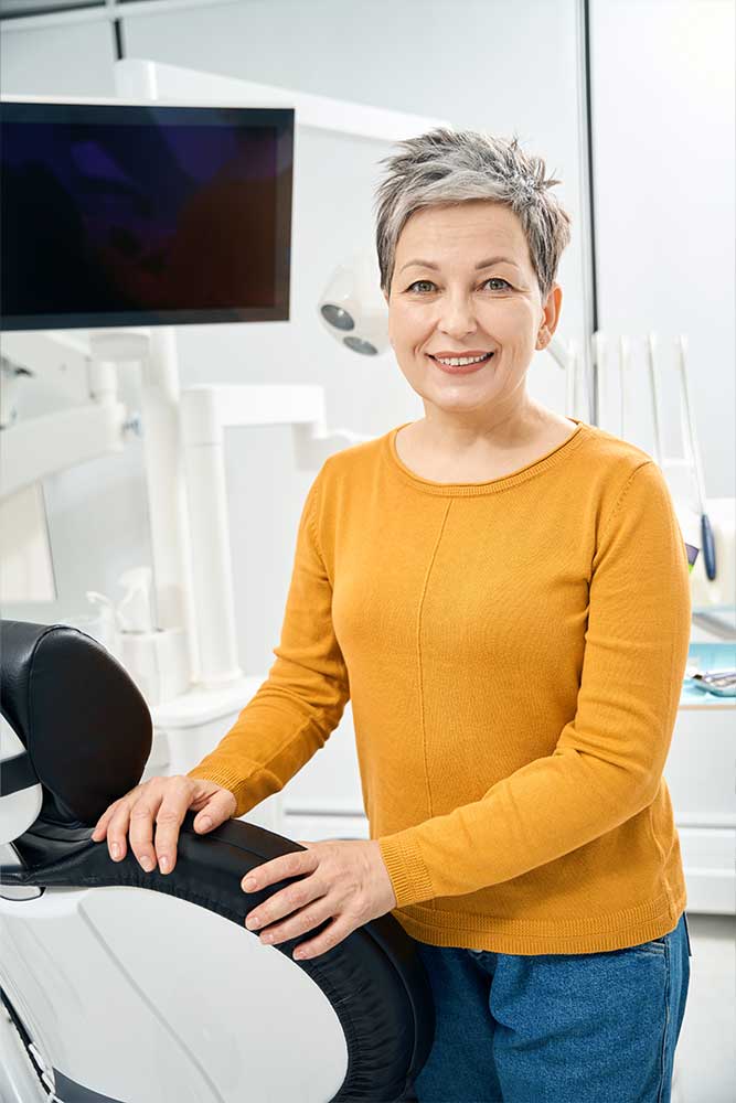older woman stands beside the dentist chair showing off her new whiter smile