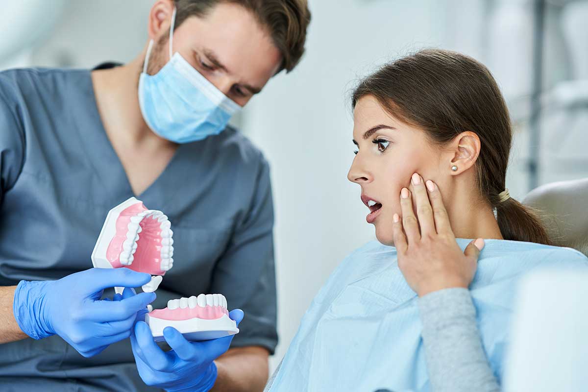 woman  who is holding her jaw in pain, is being shown how a dentist can help treat jaw pain with a plastic model of a jaw