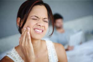 brunette woman holds her cheek as she experiences one of the common reasons for tooth pain at home with her husband computing on the couch