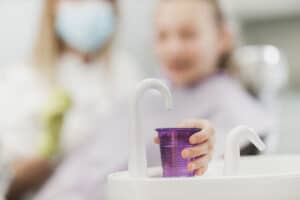 young girl at the dentist holds out a fluroide filled cup because her dentist told her about the benefits of regular fluoride treatment