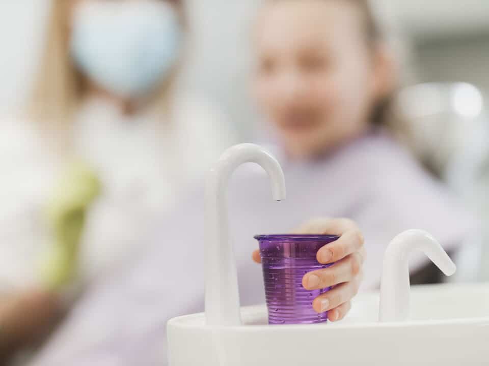 young girl at the dentist holds out a fluroide filled cup because her dentist told her about the benefits of regular fluoride treatment