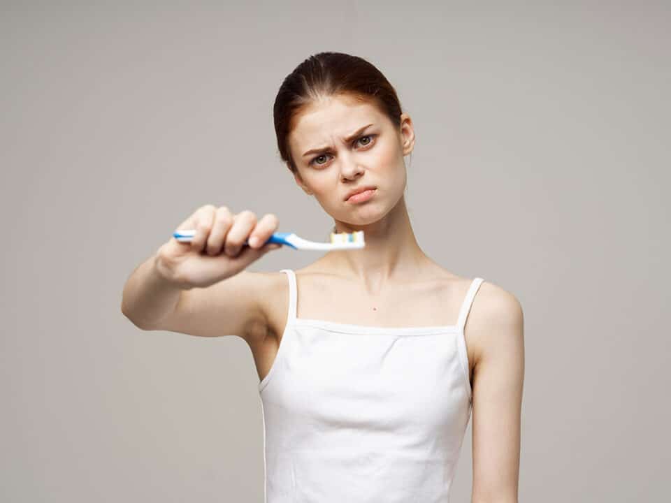 woman with a frown on her face holds out a toothbrush in front of her as she thinks about the 10 mistakes you might be making while brushing your teeth