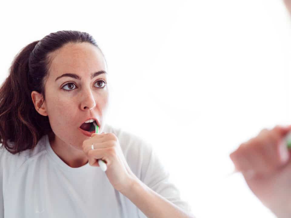 woman furiously brushing her teeth in the mirror wondering why do my gums bleed when i brush my teeth