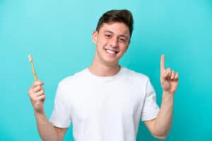young man in a white t-shirt stands in front of a blue backgrond with a toothbrush in one hand and his other finger in the air to show a way how to clean teeth without brushing
