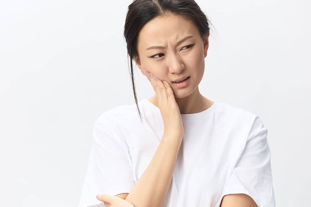 asian woman in a white tshirt holds her jaw from some painful toothache pain