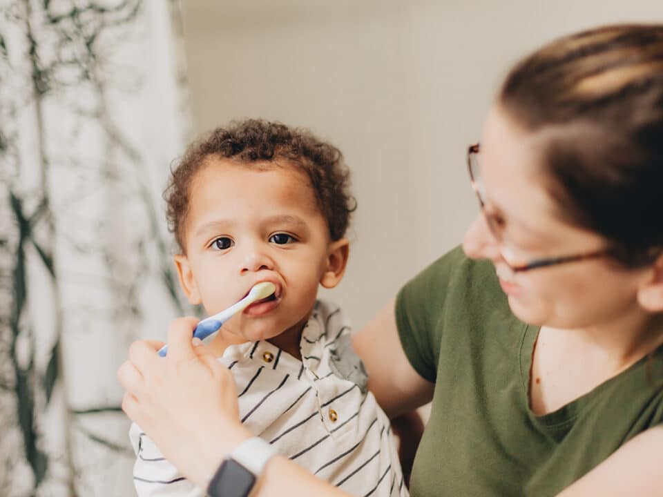 mom who knows about oral care for pediatric dental milestones is brushing her sons teeth while holding him in her arms