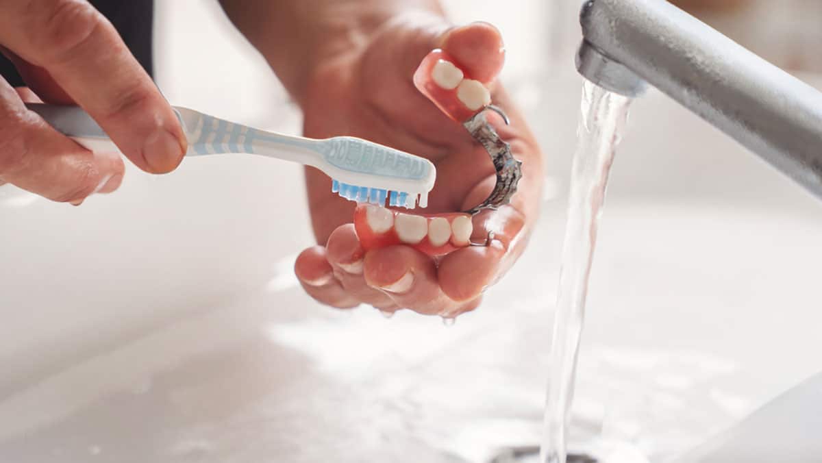 woman cleans her dentures in the sink with a soft toothbrush and running water