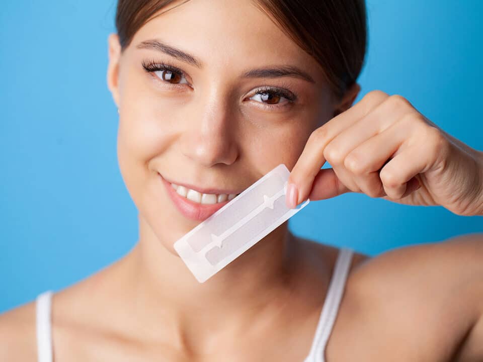 smiling woman holds up a set of strips and wonders if at-home teeth whitening strips work?