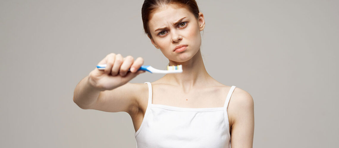 woman with a frown on her face holds out a toothbrush in front of her as she thinks about the 10 mistakes you might be making while brushing your teeth