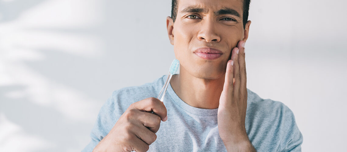young man holds a toothbrush and his cheek as he's experiencing one of the 9 common causes of tooth pain