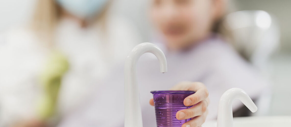 young girl at the dentist holds out a fluroide filled cup because her dentist told her about the benefits of regular fluoride treatment