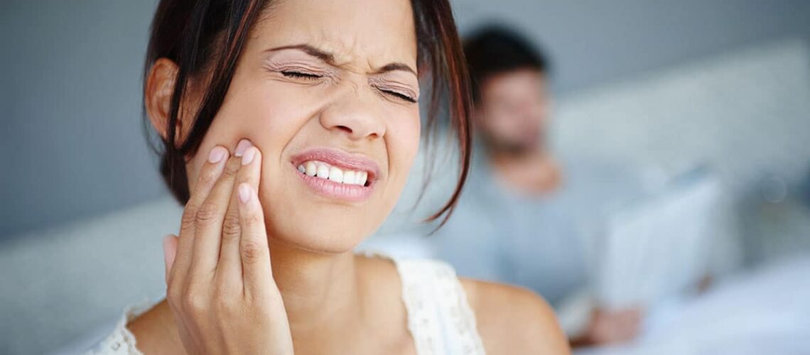 brunette woman holds her cheek as she experiences one of the common reasons for tooth pain at home with her husband computing on the couch