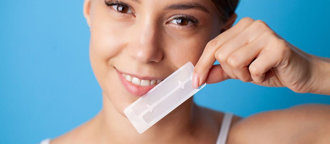 smiling woman holds up a set of strips and wonders if at-home teeth whitening strips work?