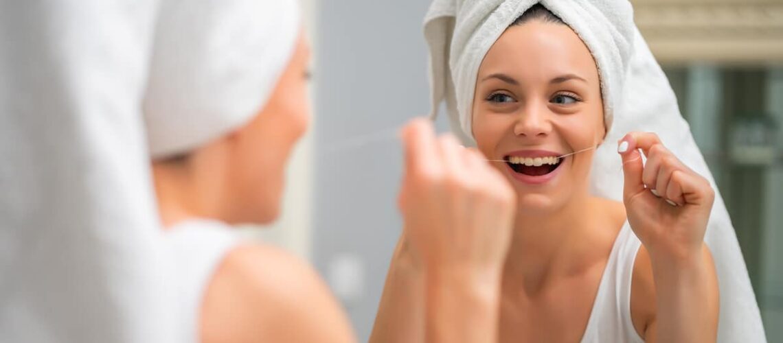 woman with her hair wrapped in a towel flosses her teeth in a mirror