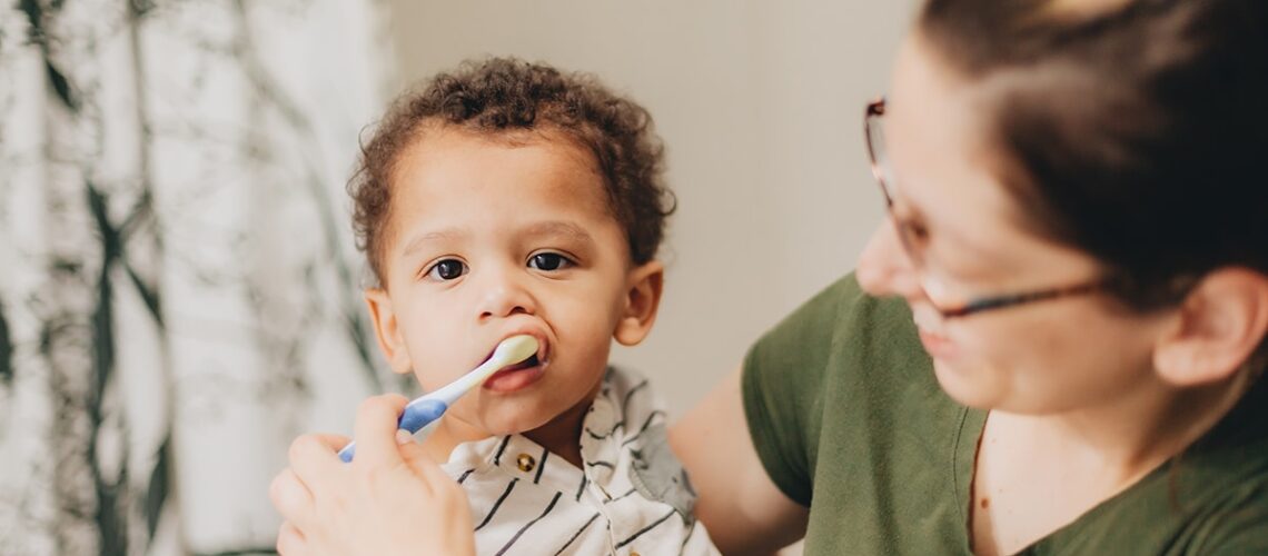 mom who knows about oral care for pediatric dental milestones is brushing her sons teeth while holding him in her arms