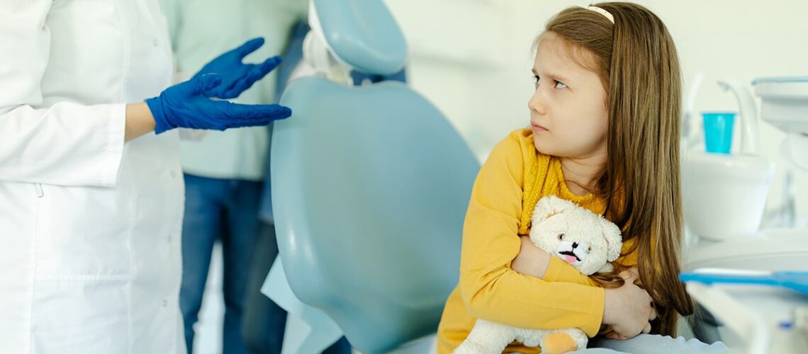 young child holds her teddy bear as she sits in the dentist chair looking frightened