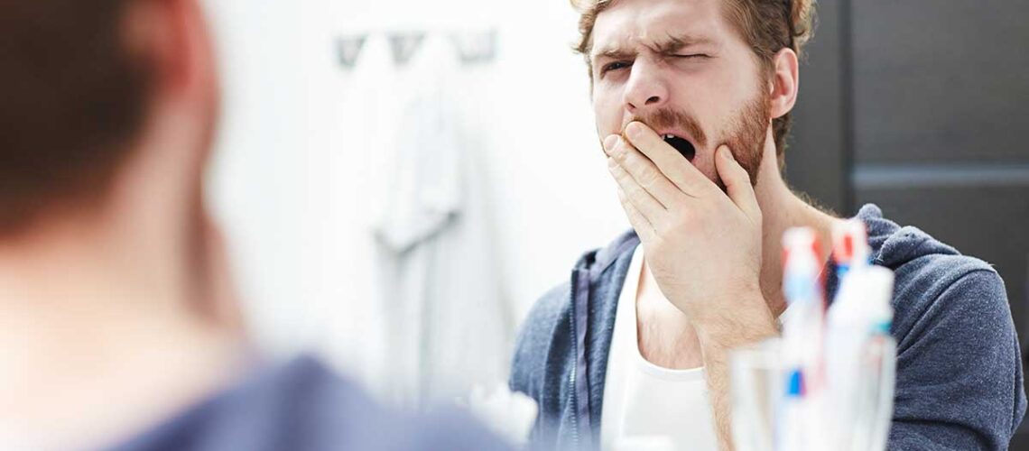 man yawning in the mirror about to brush his teeth after reading our article about tooth care for lazy people