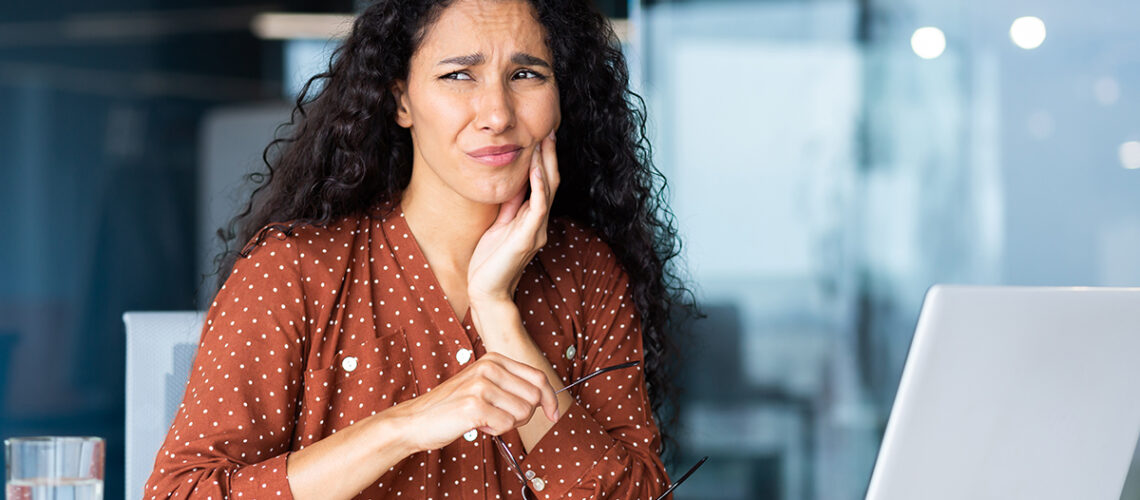 woman in red shirt sits at her desk holding her jaw as she experiences tooth sensitivity pain